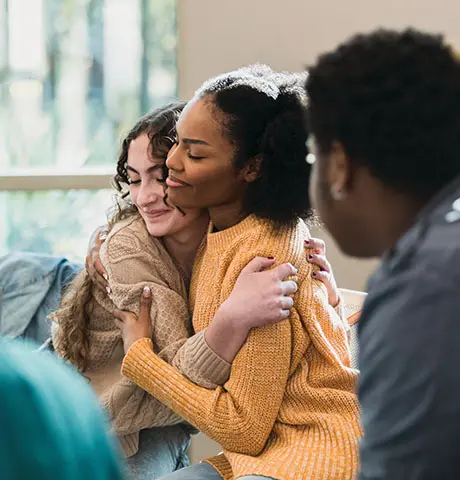 Two teen girls hug while participating in group therapy.